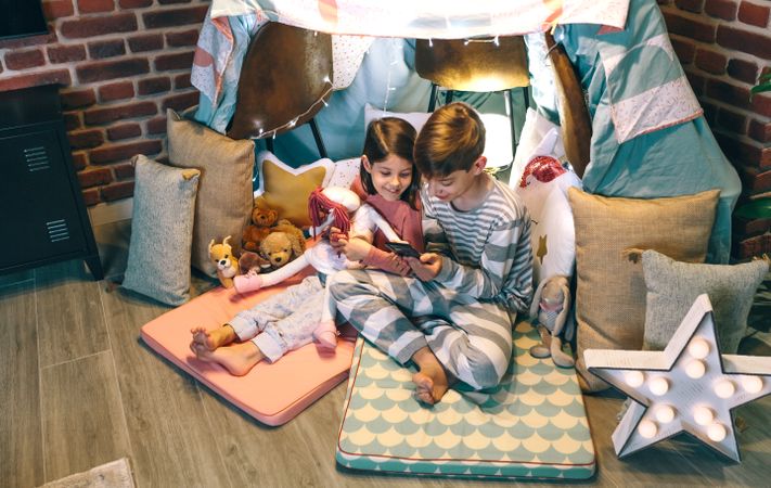 Smiling children looking mobile phone while chatting on social media application sitting on tent