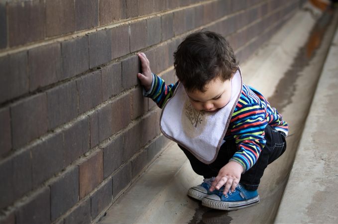 Toddler wearing onesie crouching beside gray wall outdoor