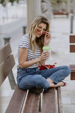 Happy woman with disposable cup of coffee sitting on street bench