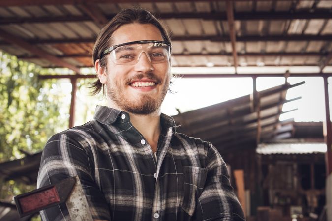 Portrait of male carpenter wearing protective glasses and holding woodworking tool in work shop