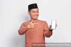 Muslim man in kufi hat smiling and pointing at blank screen on mobile phone 5QrGV4
