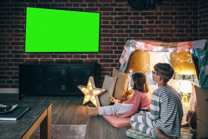 Two concentrated children sitting over mats while watching television program on living room