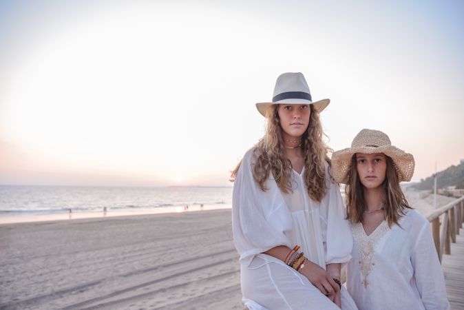 Smiling girlfriends in hats talking sitting on wooden fence on beach