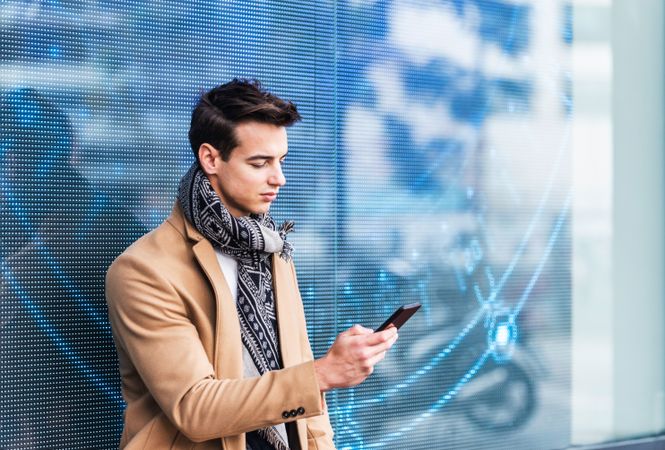 Portrait of handsome young man in fall coat against digital wall looking down at phone
