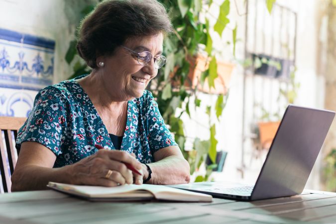 Older woman with glasses using a laptop and a notebook at home