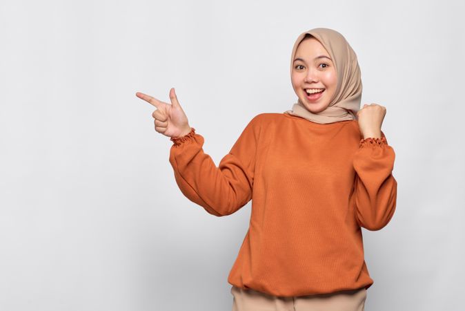 Enthusiastic Muslim woman in headscarf and orange sweater pointing finger to blank copy space