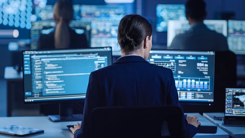 Cybersecurity Expert Monitors Network Activity on Dual Screens in Dimly Lit Command Center. Colleagues Work Nearby, Surrounded by Data-rich Displays in a High-tech Environment.