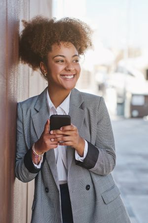 Happy Black woman using phone with smile near street wall