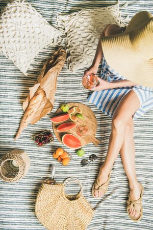 Woman on picnic blanket with  thatched bag with rose, baguette, sliced watermelon and figs