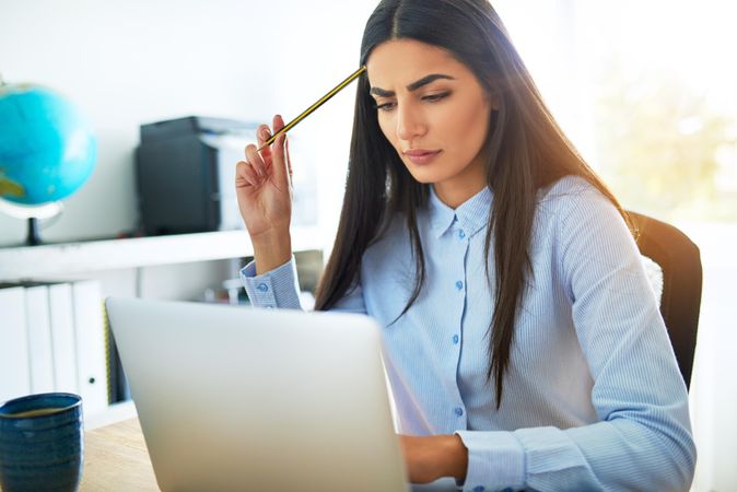 Pensive brunette woman in blue shirt working on laptop in office with pencil to head