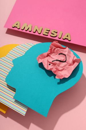 Vertical closeup of paper cut outs of colorful head on pink background with the word “Amnesia”