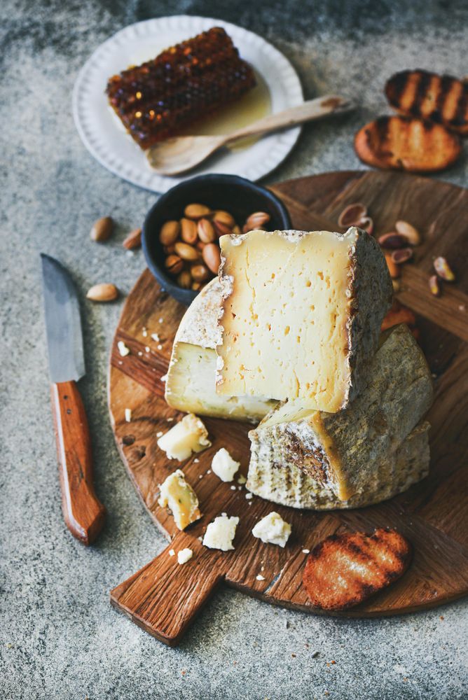 Wheel of cheese on wooden board, with knife, peanuts, crostini and honey