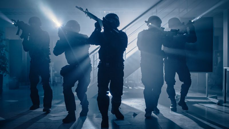 Masked Team of Armed SWAT Police Officers Move in a Hall of a Dark Seized Office Building with Desks and Computers.