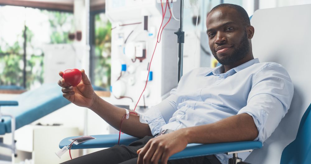 Black Man Squeezing Heart-Shaped Red Ball To Pump Blood Through The Tubing Into Bag And Showing It to Camera As A Symbol Of Support and Smiling.