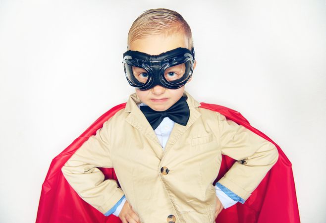 Blond boy wearing airplane goggles and cape making serious face