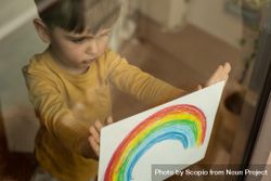 Young boy holding rainbow drawing to the window 0Vxxj4