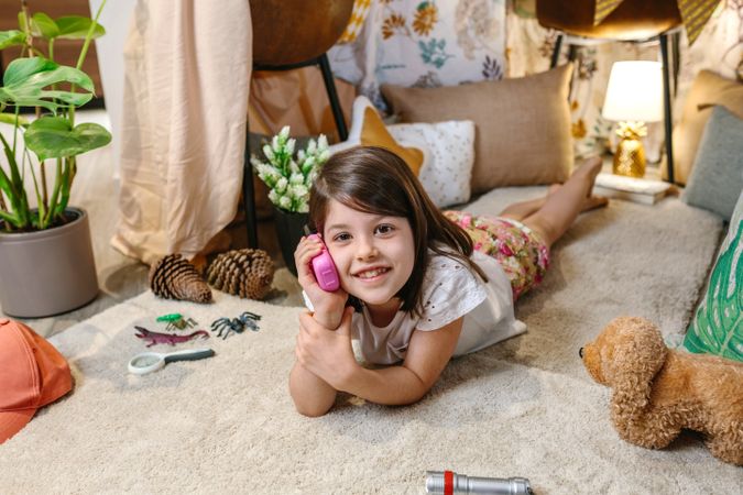 Smiling little girl looking at camera while using walkie-talkie lying over rug surrounded by cushion