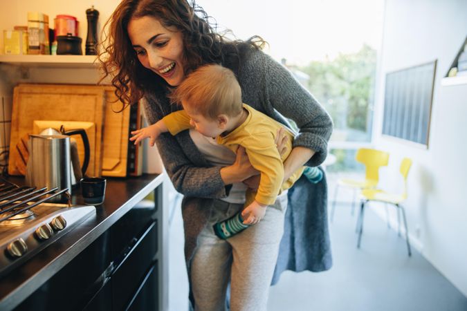 Woman carrying her son reaching out to a cup on kitchen counter