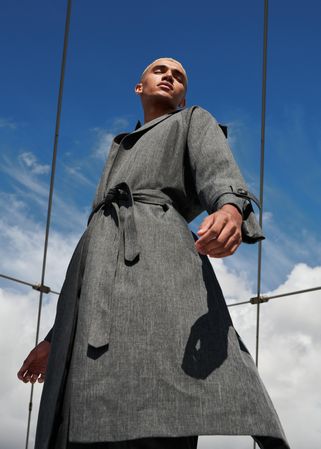 Stylish young man with eyes closed in belted gray coat against a blue sky