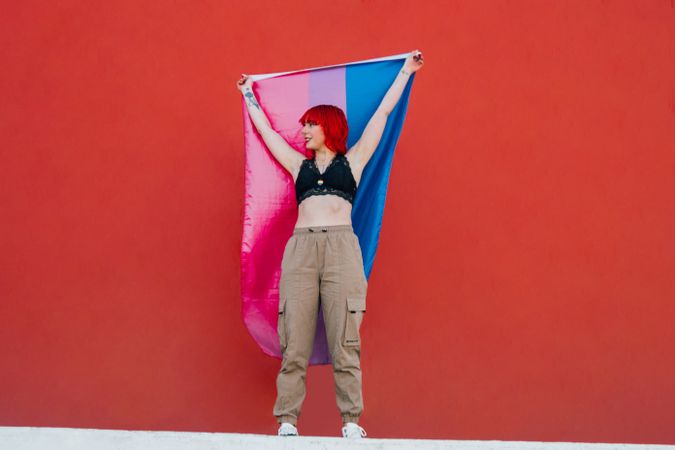 Young woman waving bisexual pride flag standing against red background