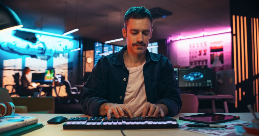 Portrait of a Young Man Typing on Keyboard and Working on Desktop Computer in a Creative Office with Neon Lights.