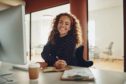 Curly haired businesswoman taking notes and smiling at her work computer