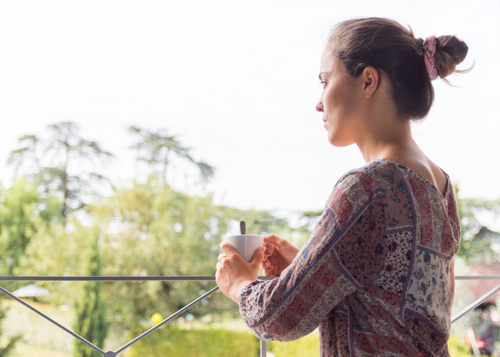 woman in a shirt with a cup of coffee or tea looking out the window in the morning