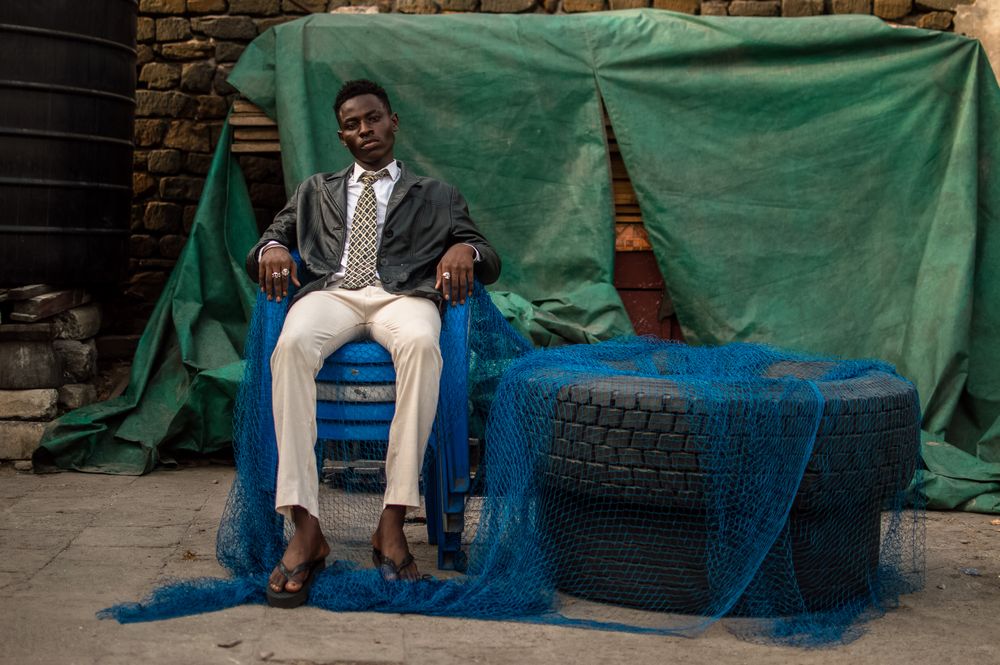 Man in dark suit jacket and tie sitting on stack of plastic chairs beside car wheels outdoor