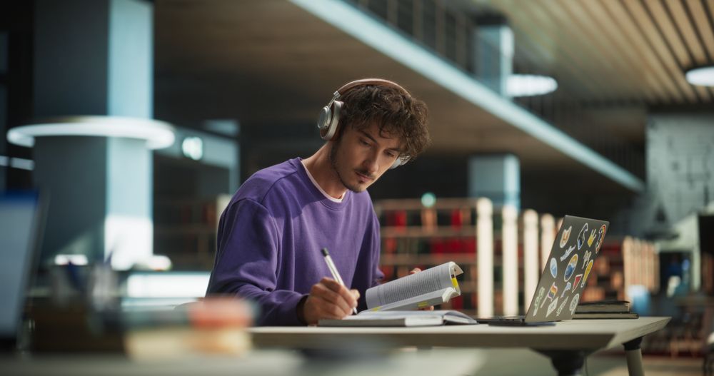 European Student Studying in a Traditional Library. Young Male Wearing Headphones, Working on an University Research Project, Reading Academic Textbook and Journals Online