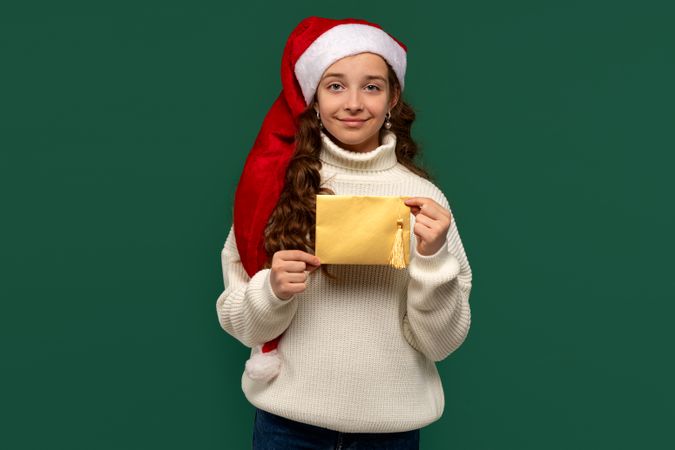 Girl in Santa hat holding golden holiday envelope.