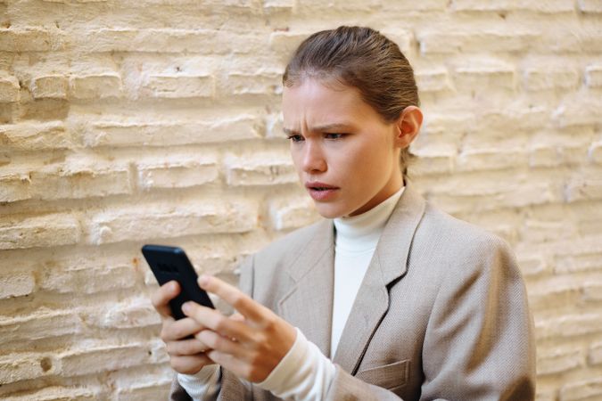 A concerned individual using a smartphone while standing against a textured wall outdoors