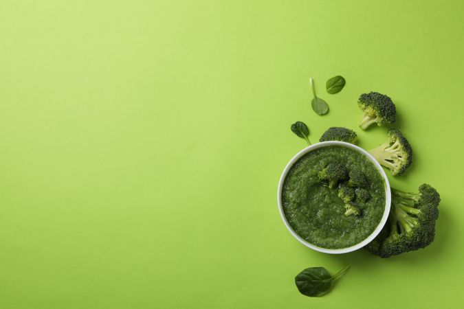 Top view of bowl of broccoli soup surrounded with vegetables on green table, copy space
