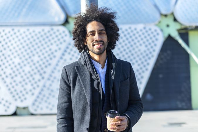 Cheerful Black man in jacket with warm beverage standing outside leaning on pole