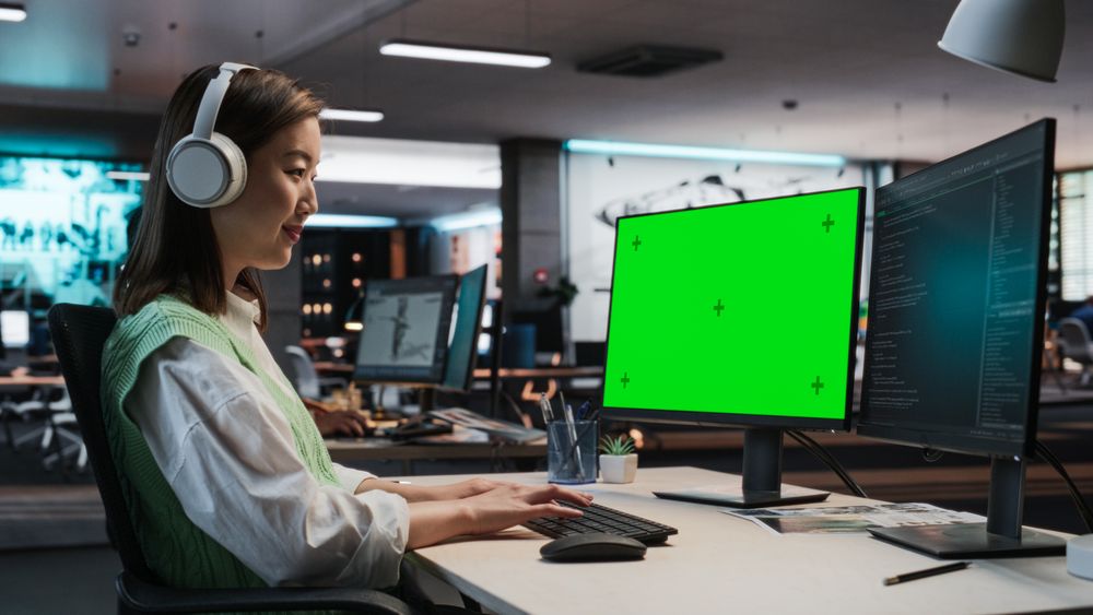 Female Asian Game Programmer Coding On Desktop Computer With Green Screen Chromakey On Display In Game Design Studio Office.