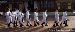 Korean men in light robe and dark hat walking in line beside building 4BE9P5