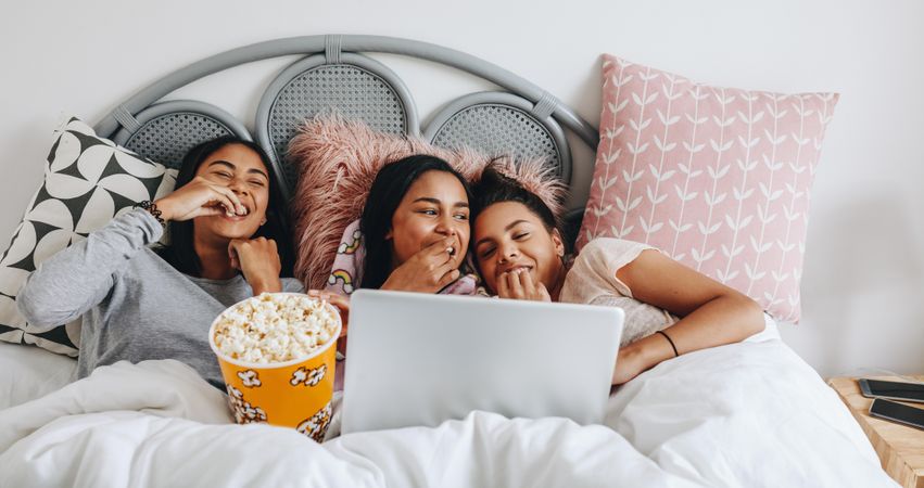 Young women having fun watching movie on laptop eating popcorn in bed