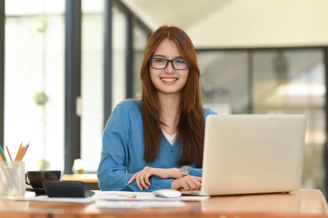 Asian woman wearing glasses and laptop on desk, looking at camera