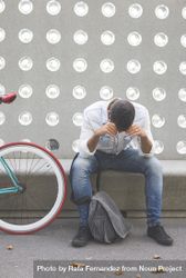 Tired male sitting with bike parked in front of patterned cement wall, vertical 0VZn30