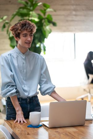 Nonbinary person standing and talking at a wooden table with laptop and coffee