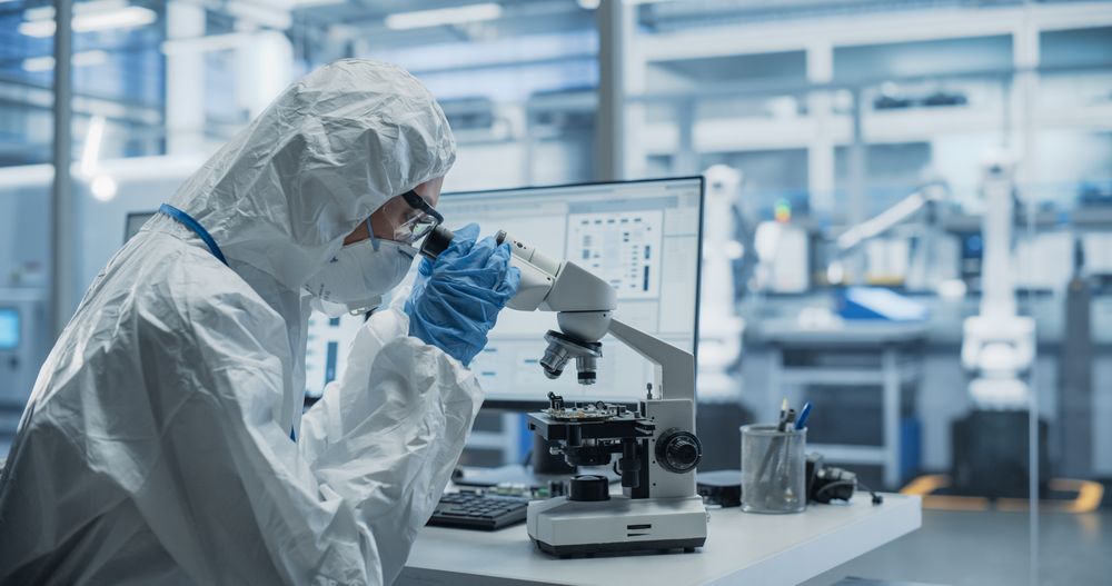 Research Factory Cleanroom: Engineer Wearing Coveralls, Gloves and Goggles while Using Microscope to Inspect a Microchip. Scientist Developing an Advanced Electronics Component for Precision Industry