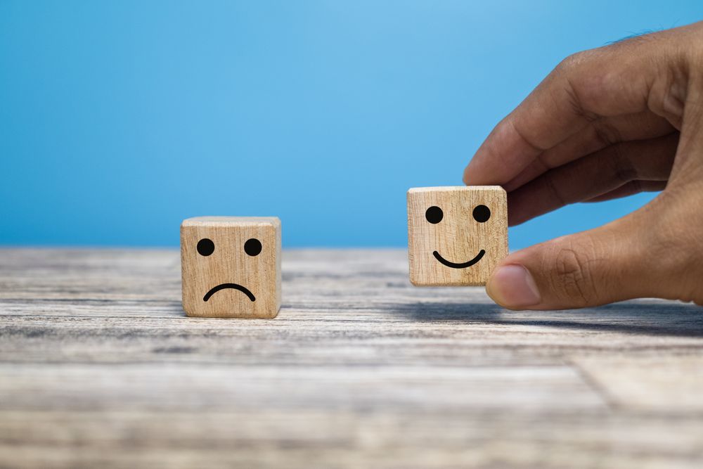 male hand putting happy face and sad face expression emoji on wooden block isolated on blue background