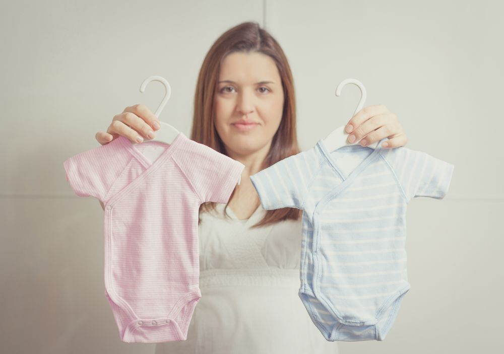 Pregnant woman holding bodysuits  for a girl or a boy in soft pastel colors