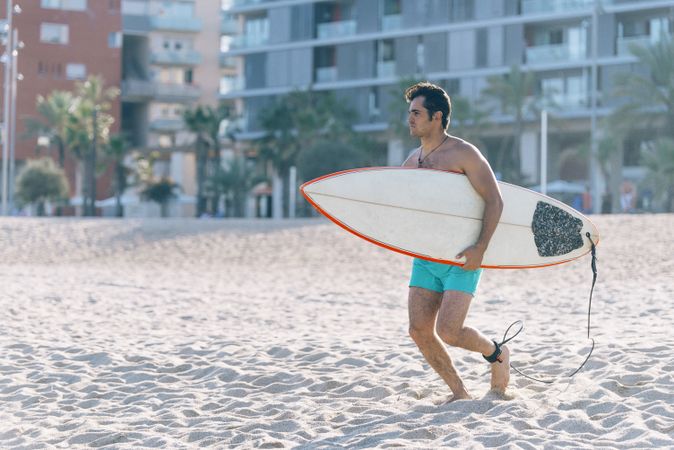Tanned male surfer walking on beach with board attached to his leg with leash