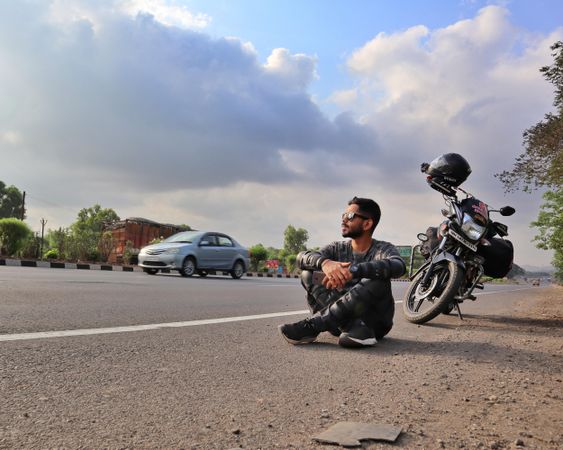 Young man sitting beside motorcycle on road