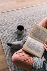 Cropped image of a person wearing socks reading a book and sitting on rug beside cup of tea 0gqaM4