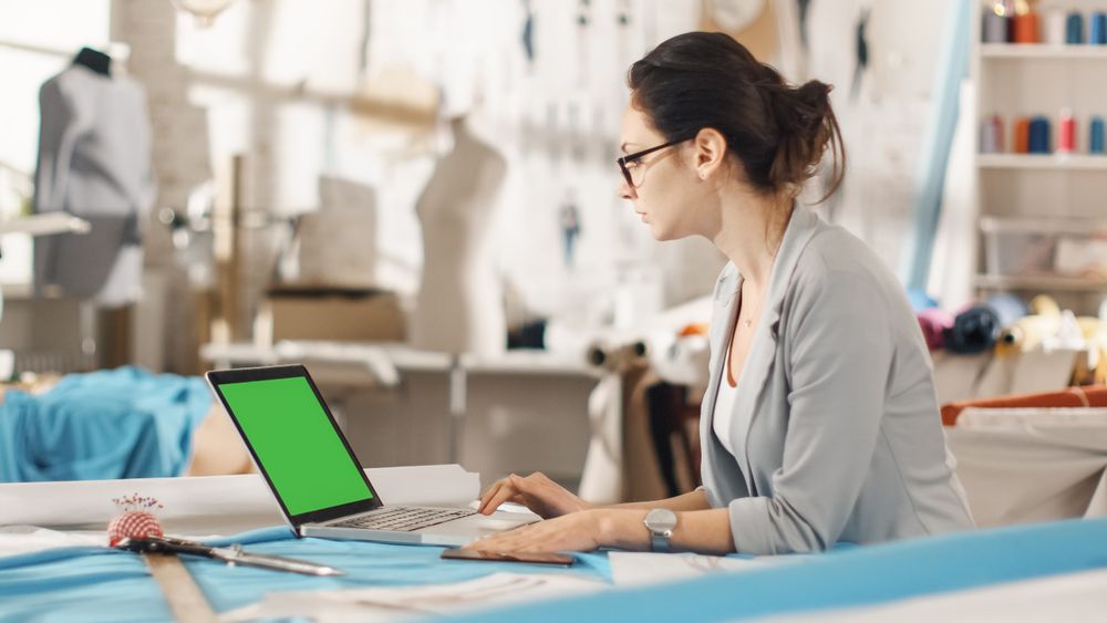Woman fashion, designer,  Sitting at Her Desk Working on Laptop with Green Screen, Taking Notes. Her Studio is Sunny,