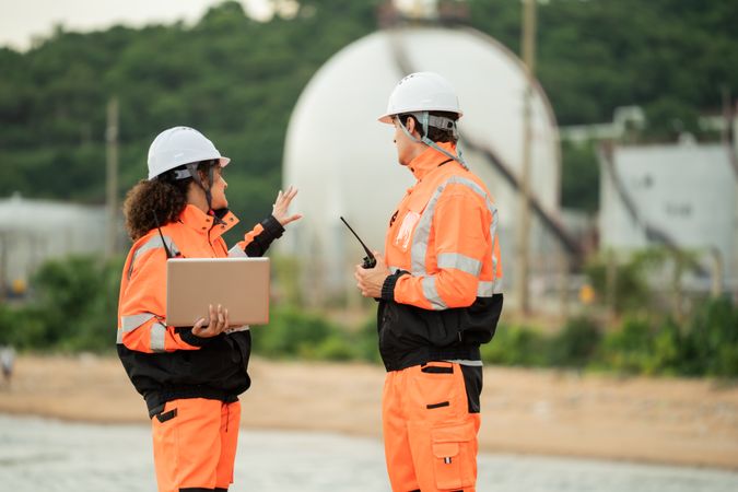 Two engineers in safety gear talking work near spherical gas storage tanks. woman is pointing forward while the man checks a laptop, indicating active discussion and site coordination in an industrial