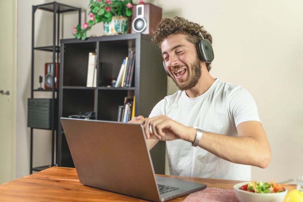 Young man laughing out loud while using a laptop with headphones at home.