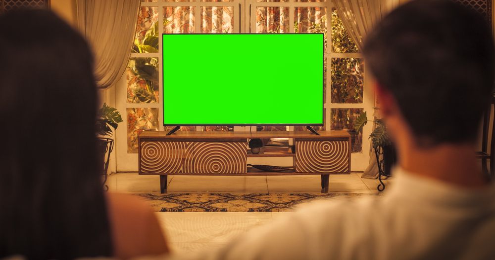 Back View of Young Indian Couple Watching Green Mock-up Screen Chroma Key TV while Sitting on a Couch in the Living Room.