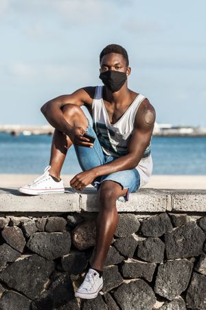 Man with facemask holding a phone sitting beside seashore